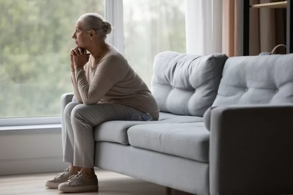 Side view of older woman sitting on gray sofa gazing pensively out of wall-sized window.